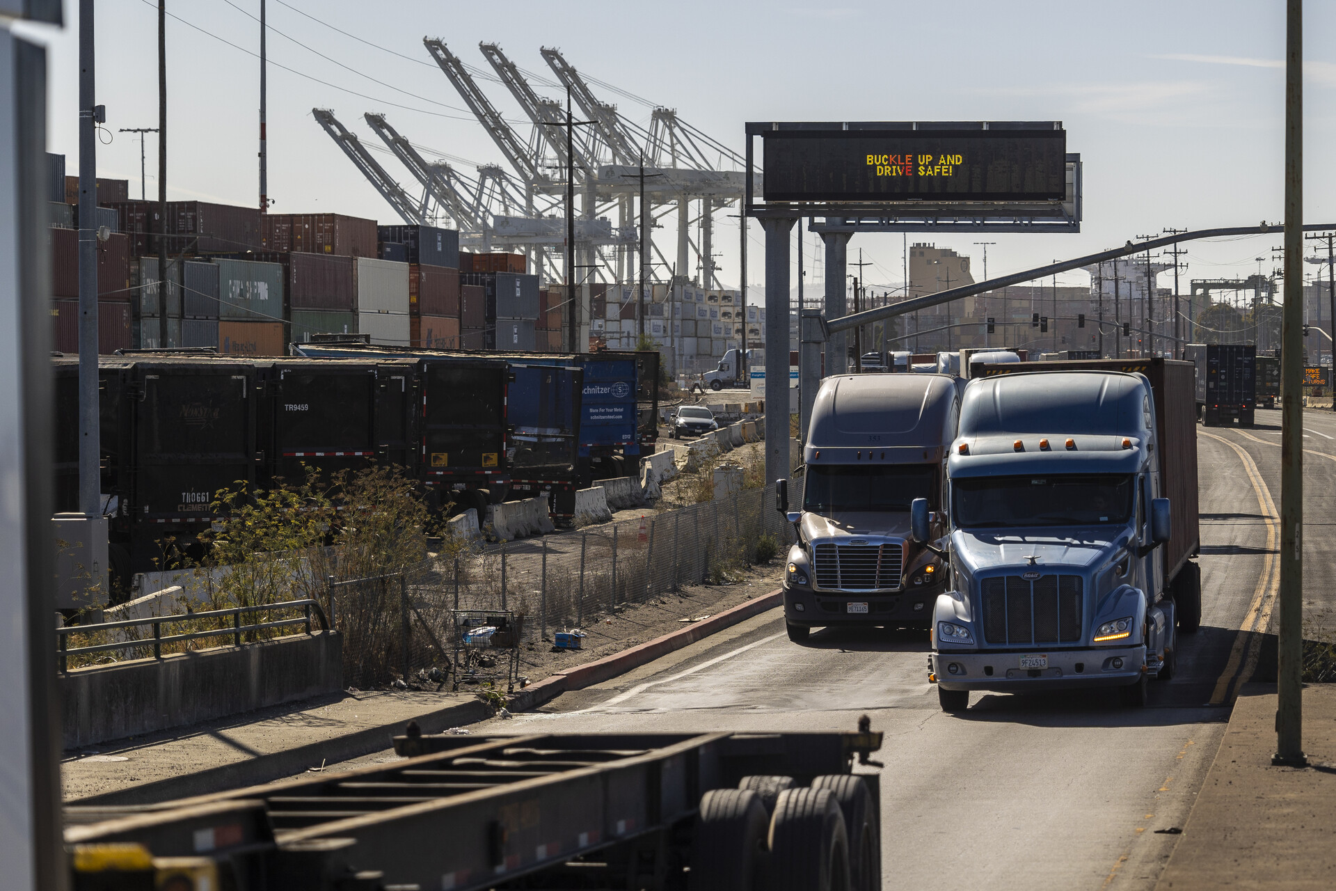Large trucks leave an industrial site, with cranes in the background.