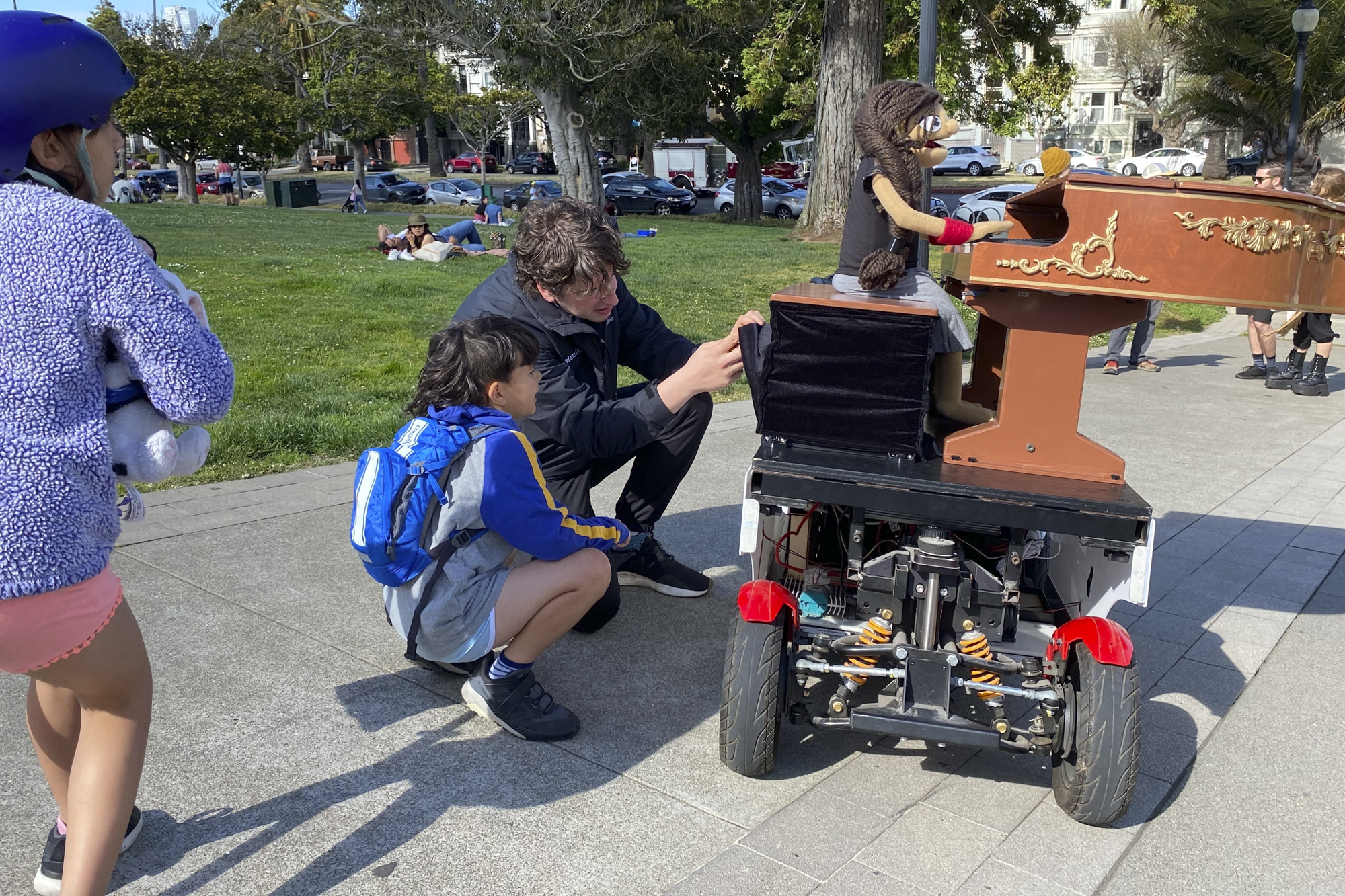 An adult and a child examine a machine atop which sits a puppet playing the piano in an outdoor setting.