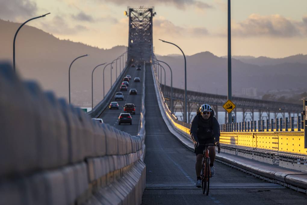A person bikes across a large bridge on which cars are also driving.