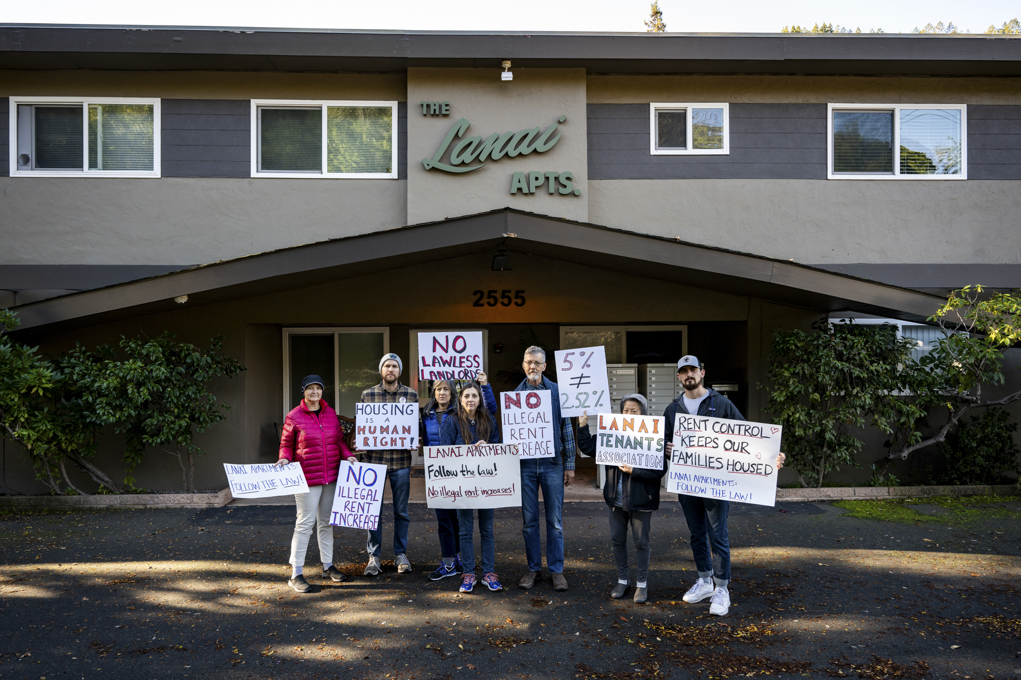 A group of seven people stand in front of a two story building holding signs and looking at the camera.