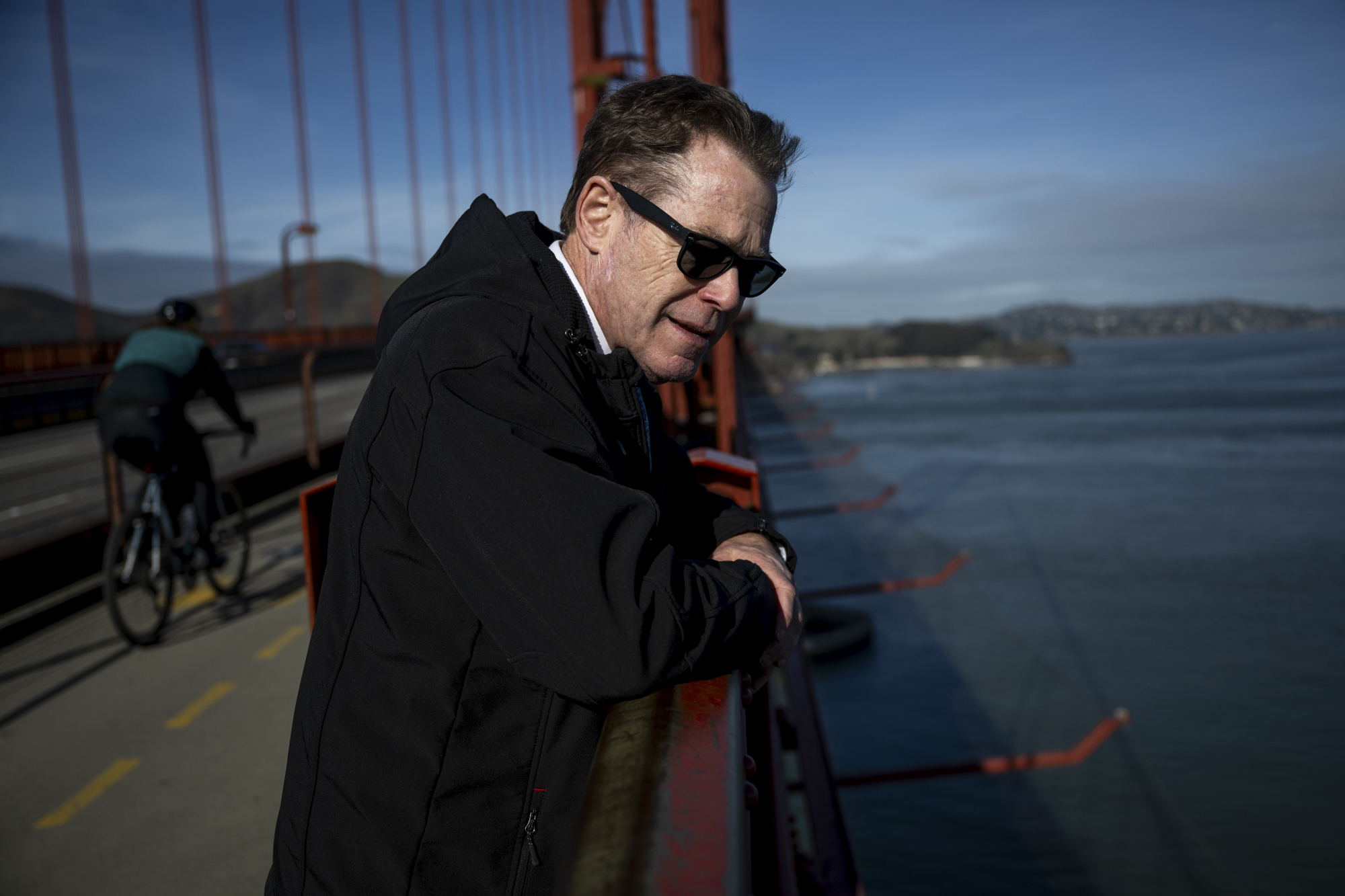 Um homem branco com terno e óculos de sol olha para a beira da ponte Golden Gate.
