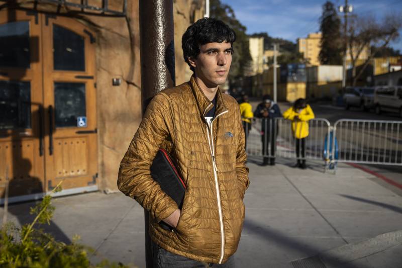 A person in a light brown jacket stands on a street corner.