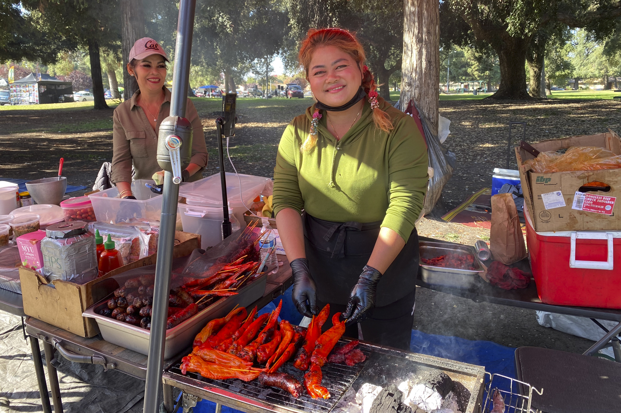 A person with long hair smiles while tending to a grill in a park setting.