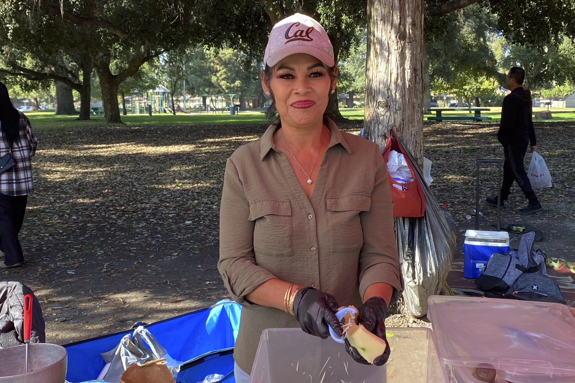 A person wearing a baseball cap smiles while working with food in an park setting.
