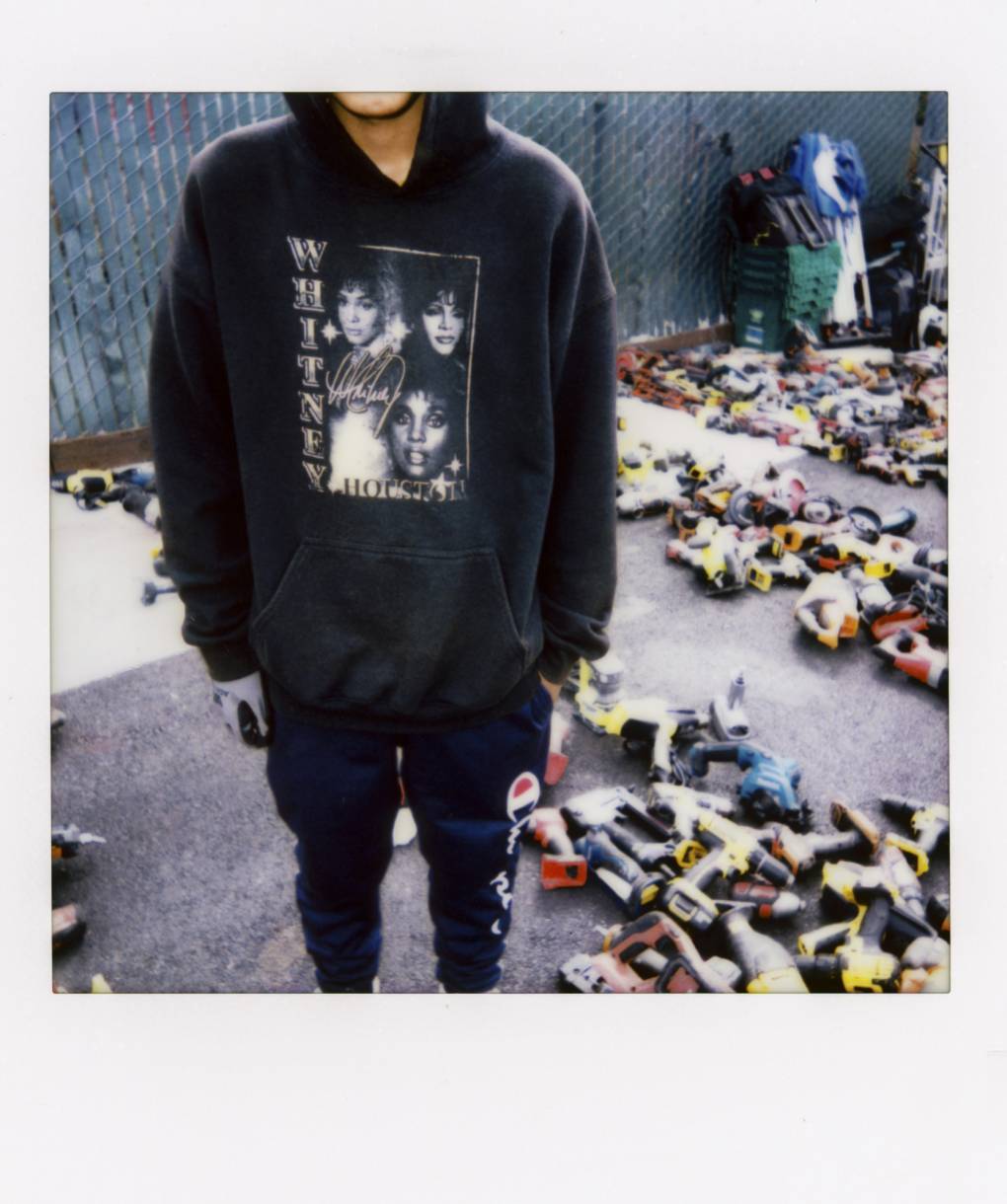 A person wears a Whitney Houston sweatshirt while standing beside an array of power tools in an outdoor setting.
