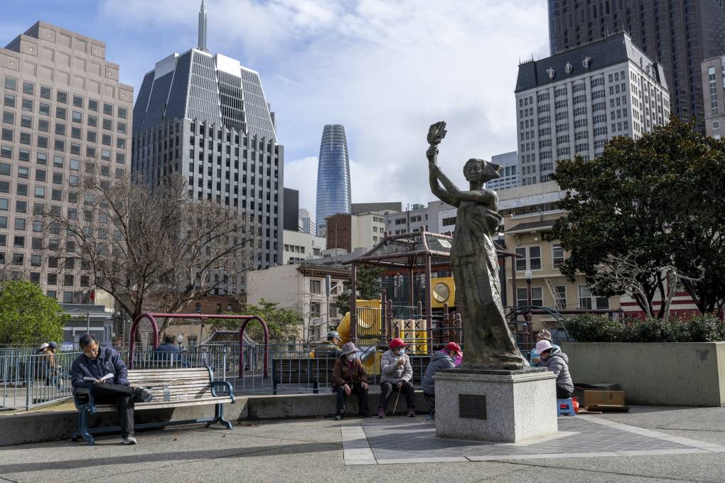 People sit on a bench with a statue in front of them. Tall buildings surround them in the background.