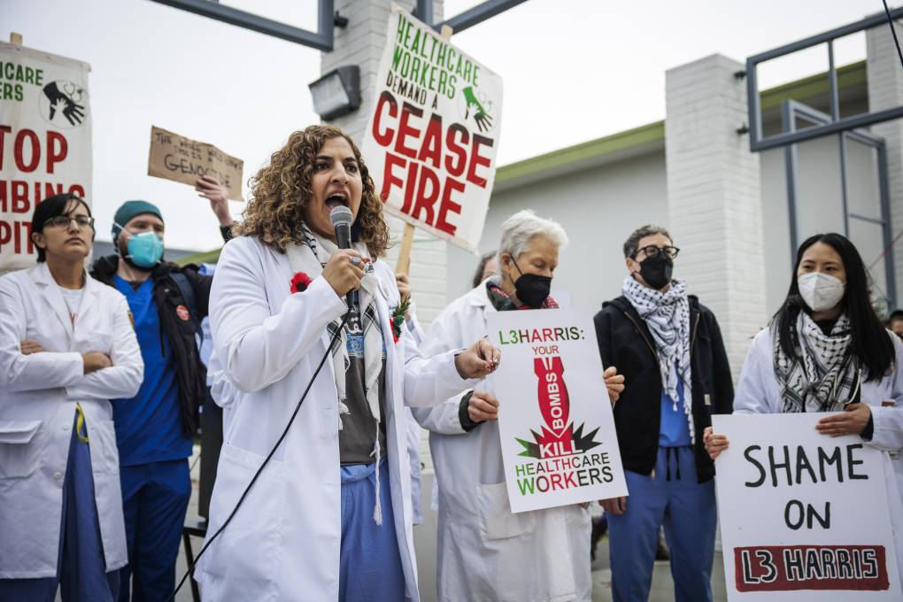 A woman wearing a labcoat talks into a microphone at a rally of health care workers also wearing labcoats holding posters calling for a cease-fire in Gaza.