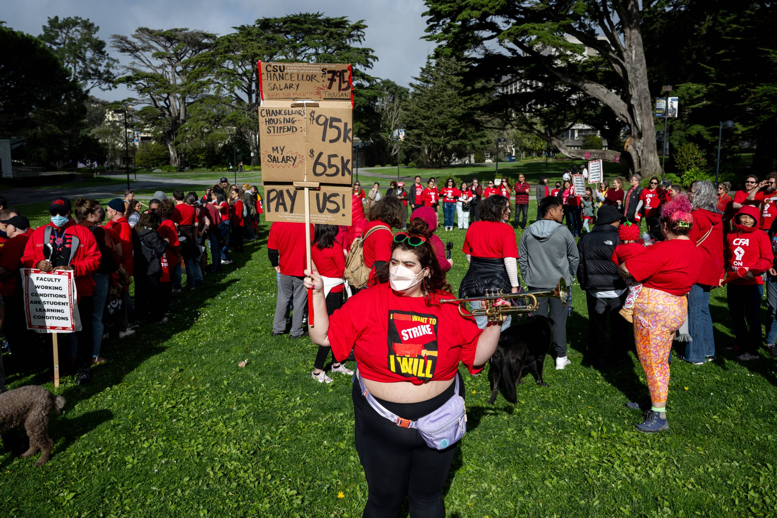 A person wearing a red t-shirt holds a cardboard sign in one hand and a trumpet in the other while standing on a grassy area in front of a group of people in mostly red attire.