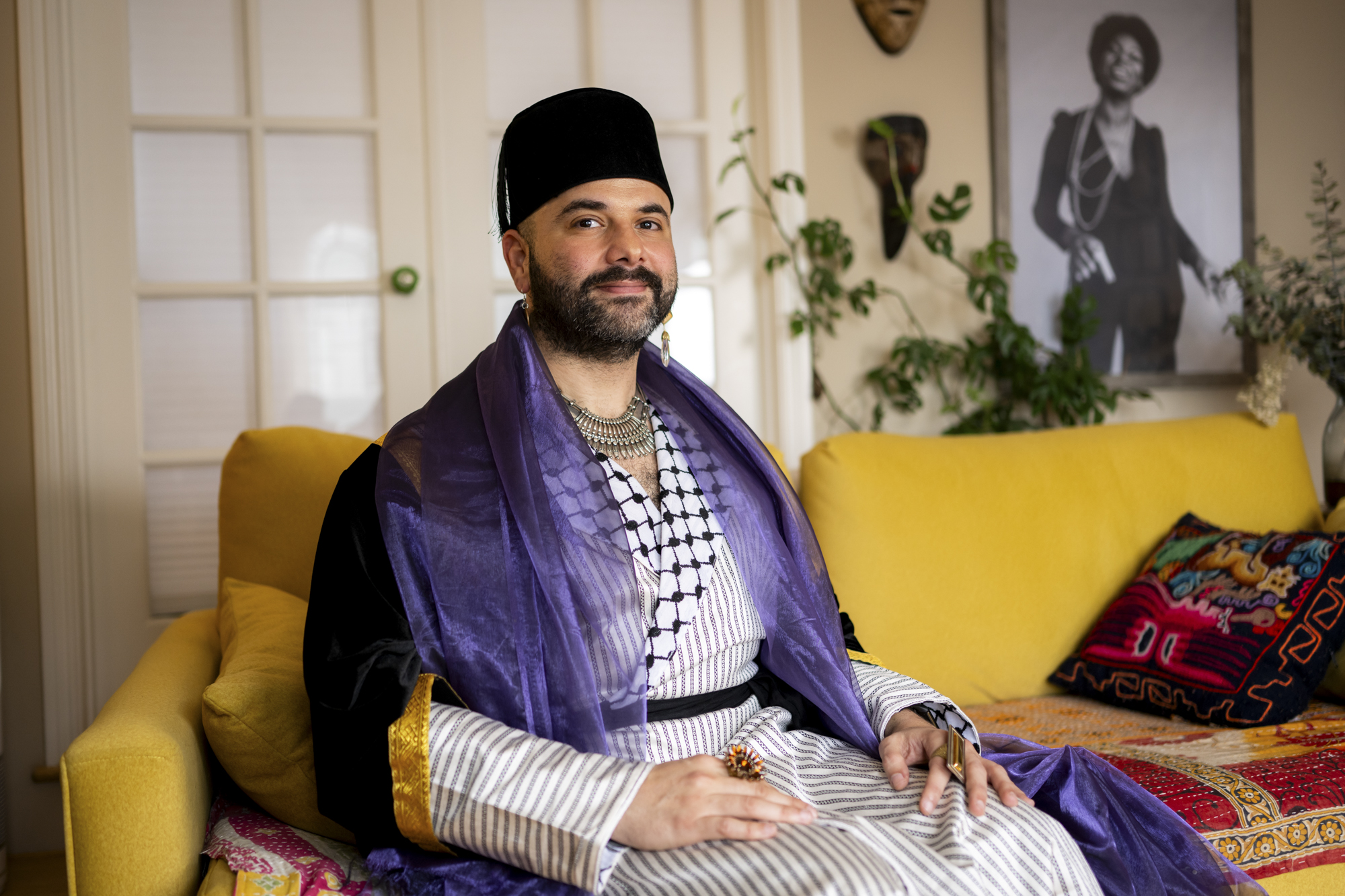 A person with a beard wears a hat and sits on a sofa smiling and looking at the camera.