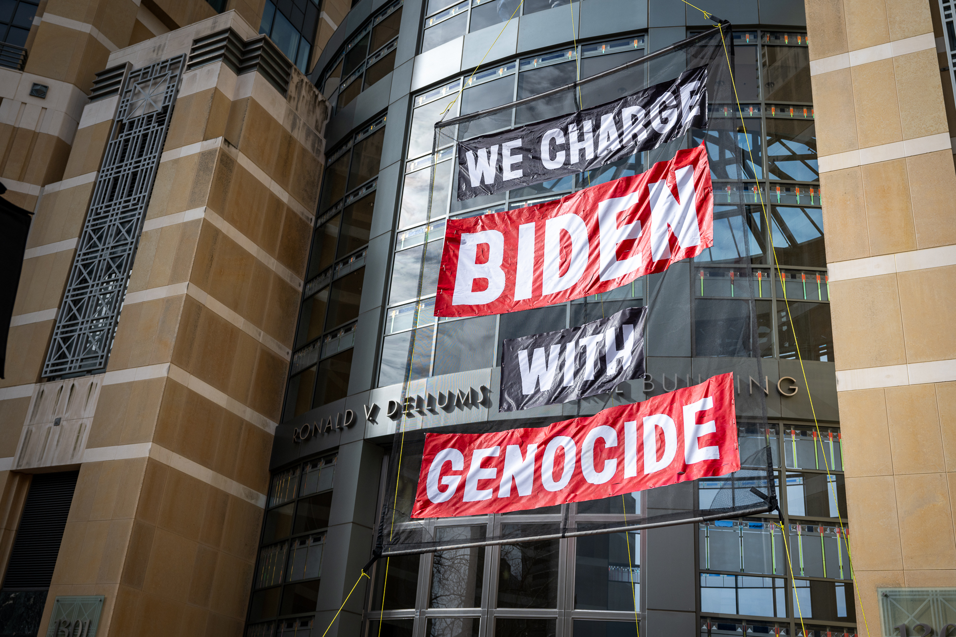a large sign hangs from a courthouse building that reads 'we charge Biden with genocide'