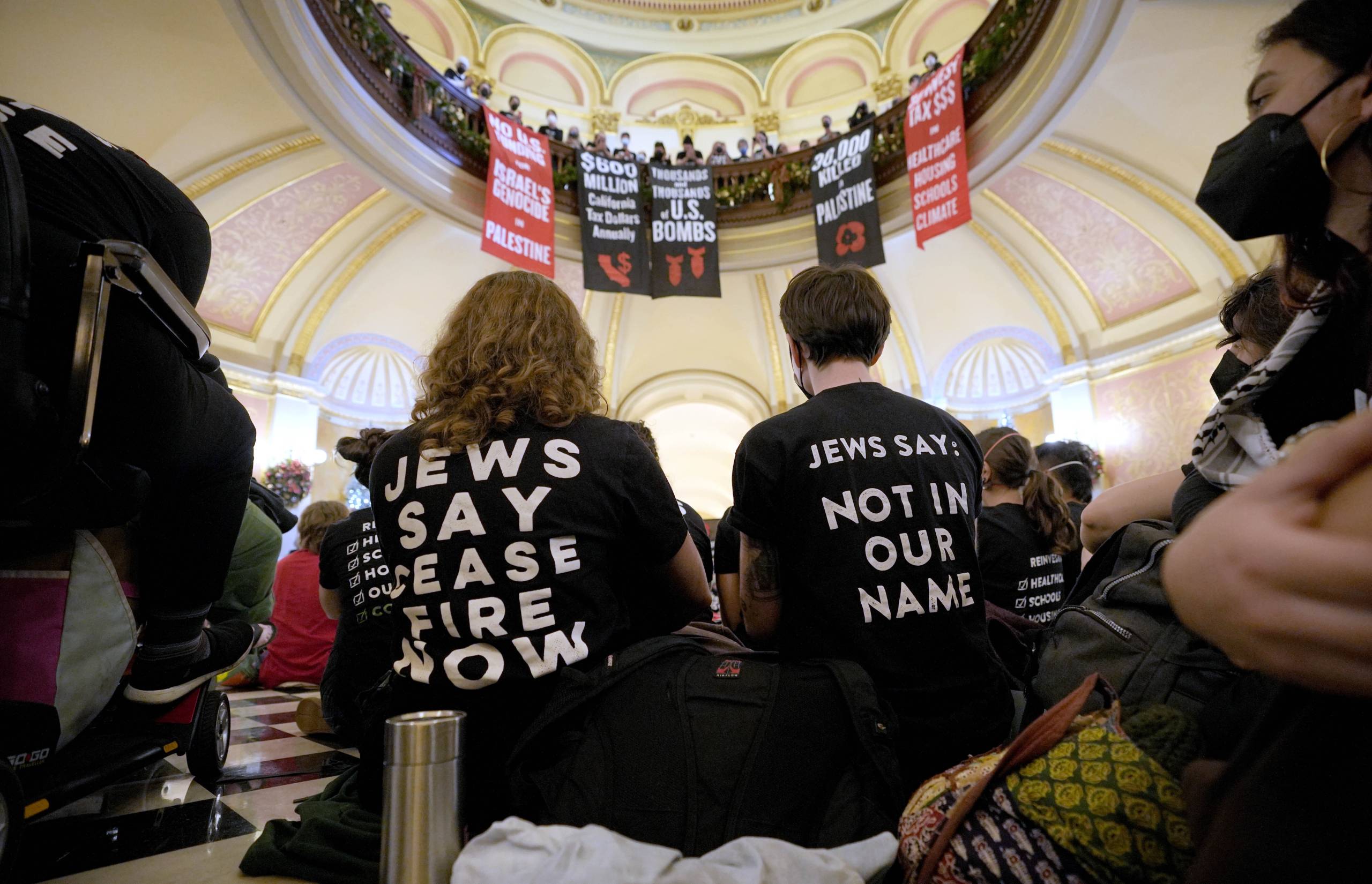 Two people with backs turned to the camera look up at signs advocating for a cease-fire with the background of the California state capitol.