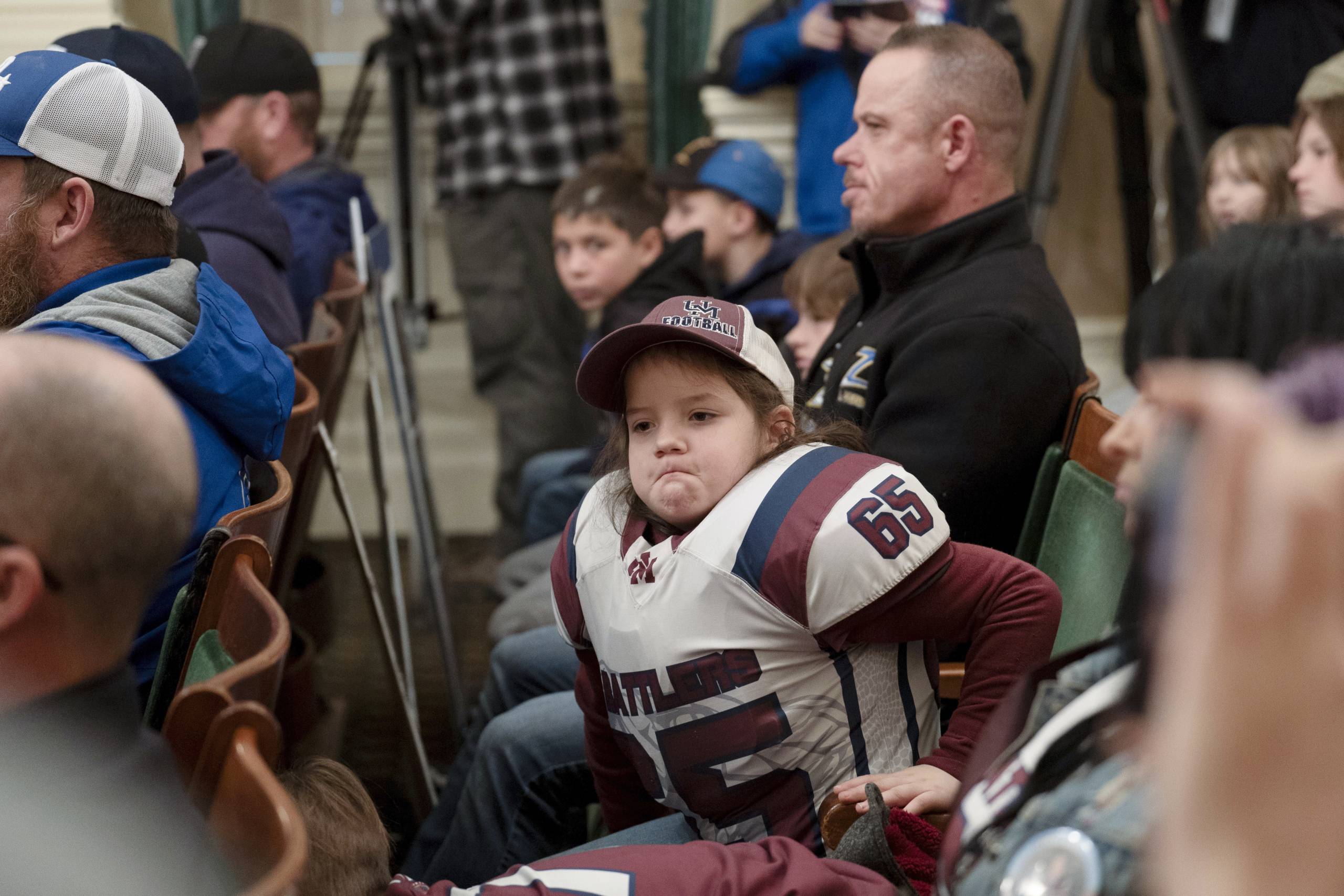 A boy wearing a football uniform and equipment sits in a chair and looks over, surrounded by other people in the auditorium.