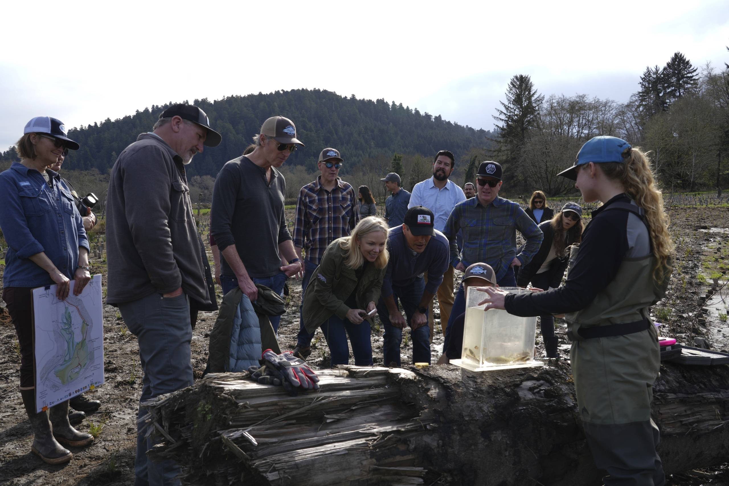 A group of people standing outside in a forest look at a fish in a container.
