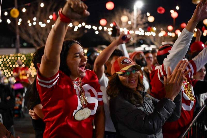 Two women wearing San Francisco 49ers paraphernalia celebrate among a larger crowd of people.