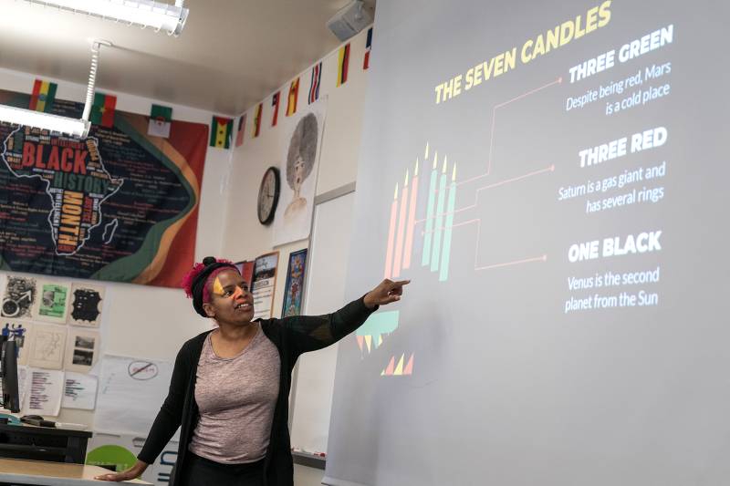 A college professor stands in front of her classroom, pointing to a PowerPoint presentation.