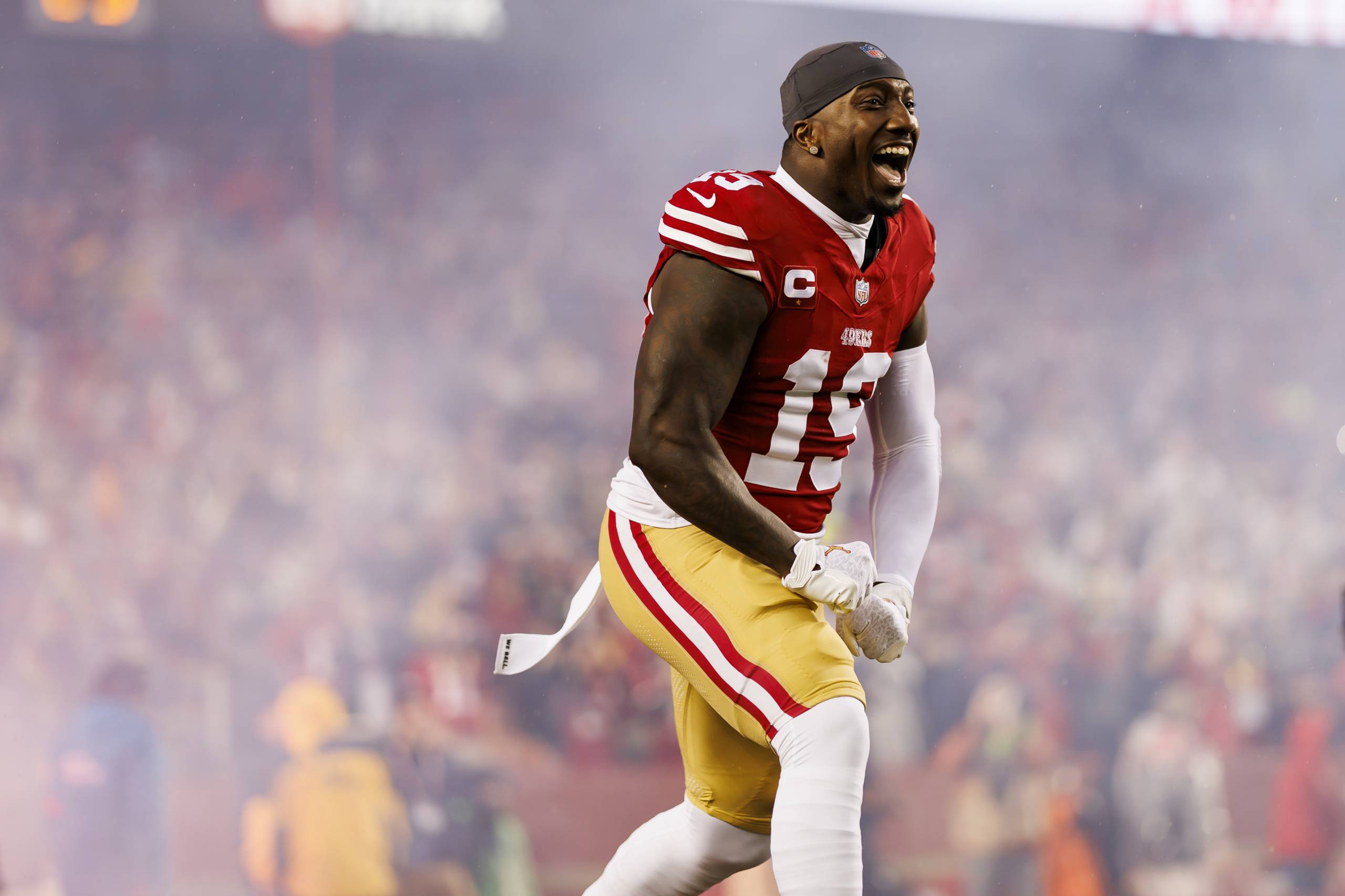 A tall, muscular man dressed in 49ers uniform stands in front of a crowd, looking away from the camera, and cheers.