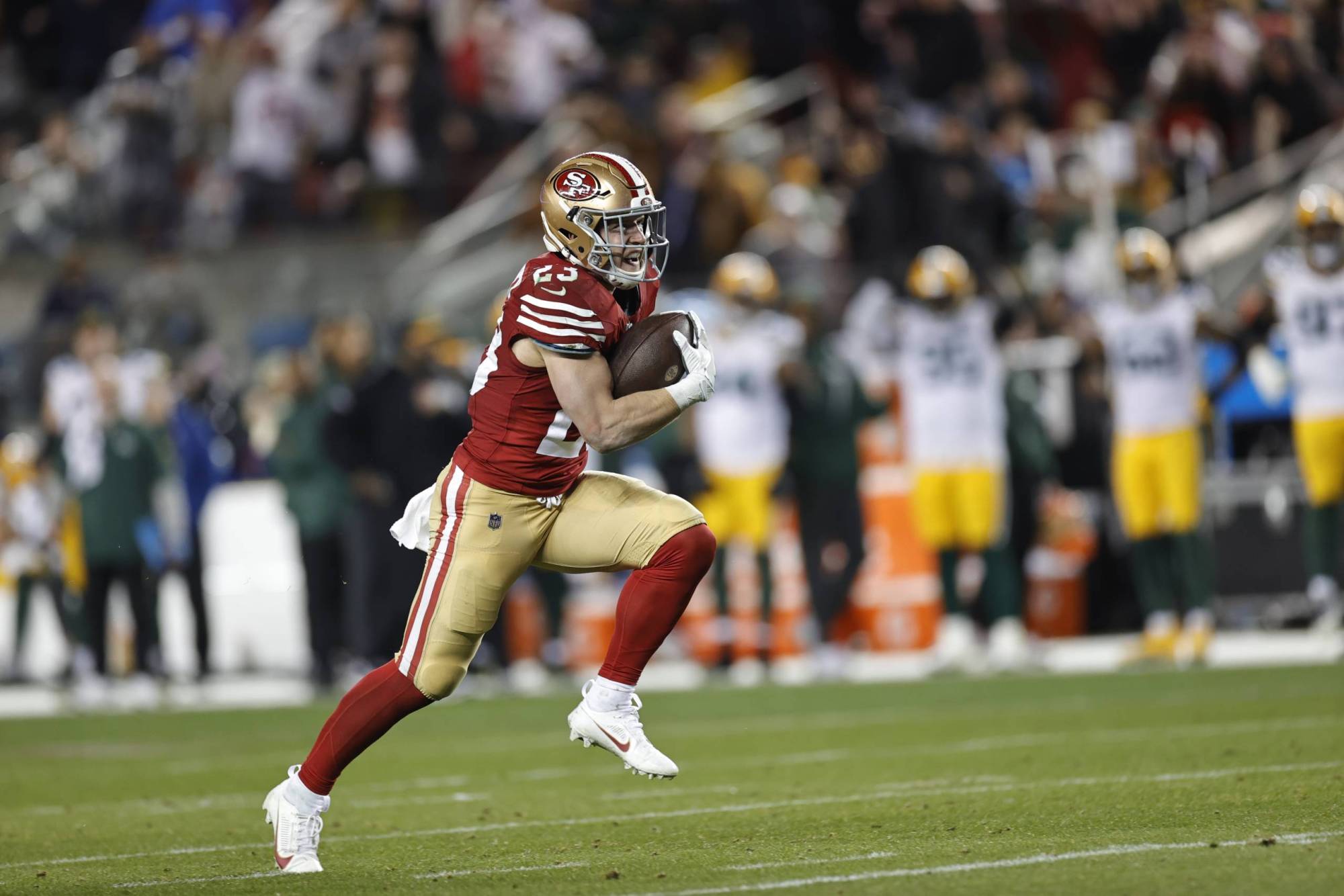 A man geared up in football uniform runs down a large football field, holding a football with his right hand.