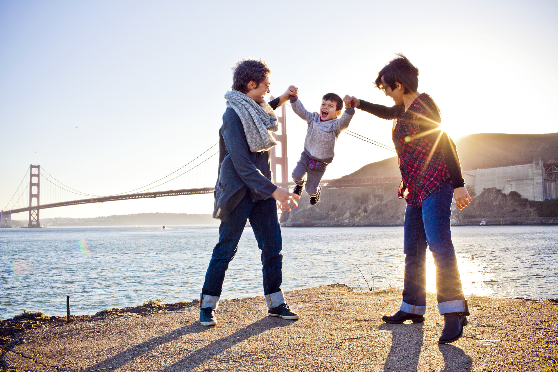 Two people swing a child by the arms in front of the Golden Gate Bridge as the sun goes down.