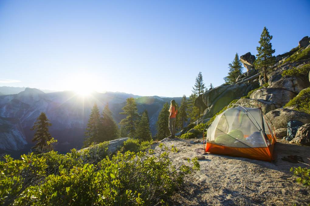 A woman looking out at the sunrise surrounded by mountains, with a tent behind her.