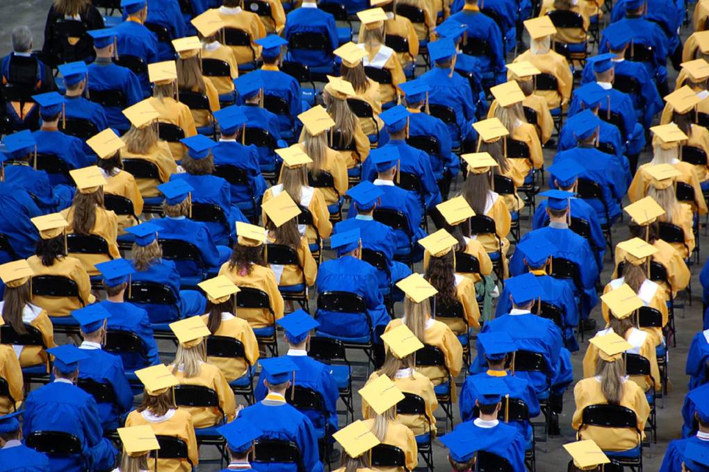 A tight shot captured from behind of hundreds of students in blue and yellow graduation caps sitting down.