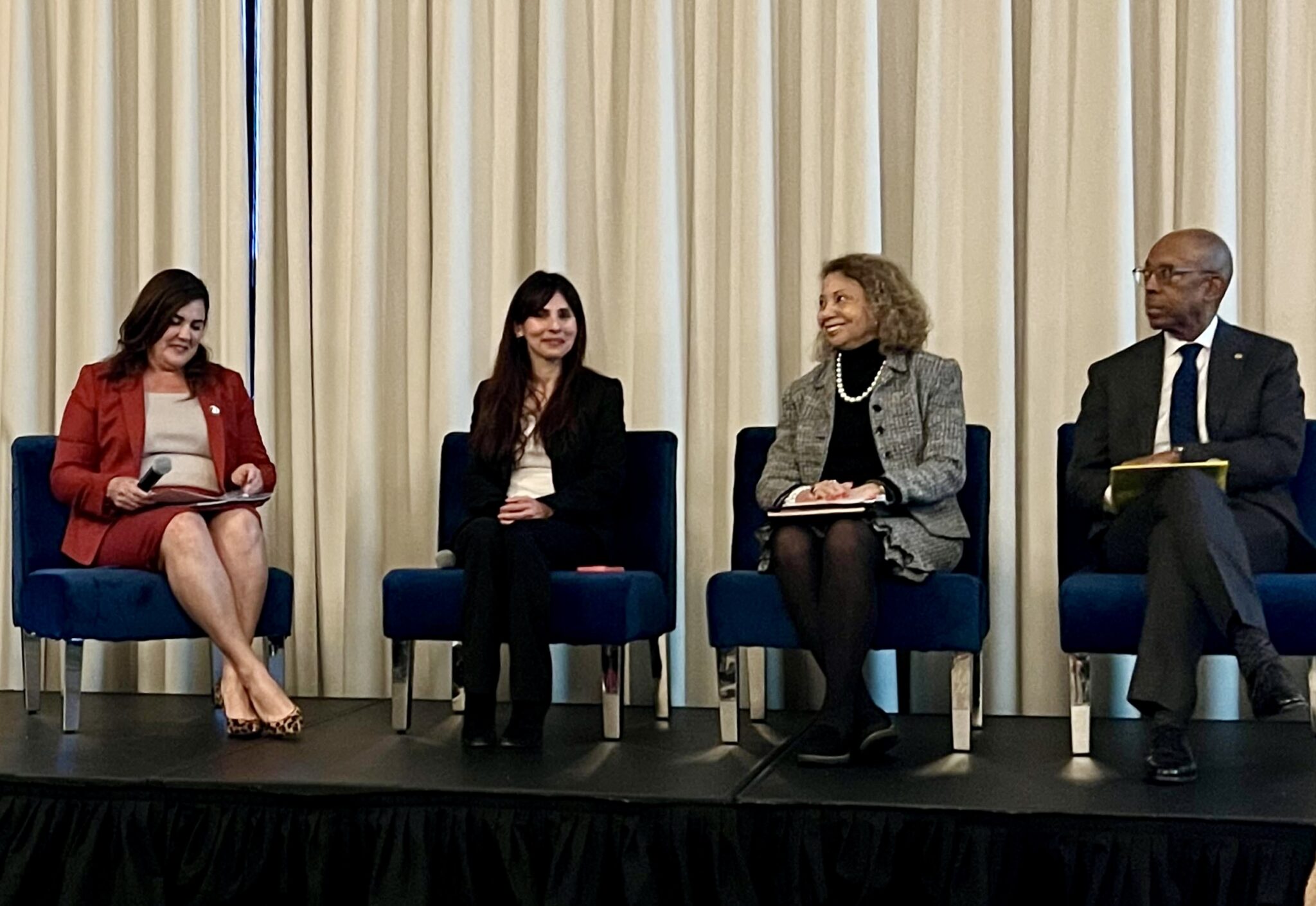 Three women and one man wearing business suits sit on a stage, looking at each other.