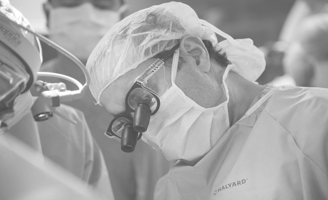 A black and white photo of a surgeon in surgeon's scrubs is seen operating with a team behind him watching his work.