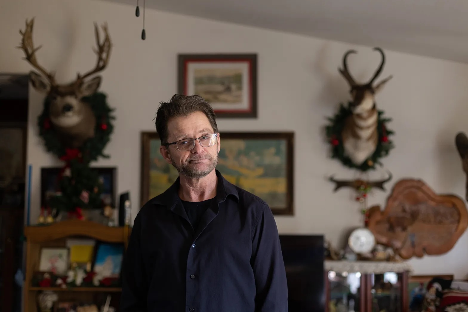 A white middle aged man looks and smiles wryly at the camera, wearing glasses, in a room with maps and deer heads mounted on the wall.