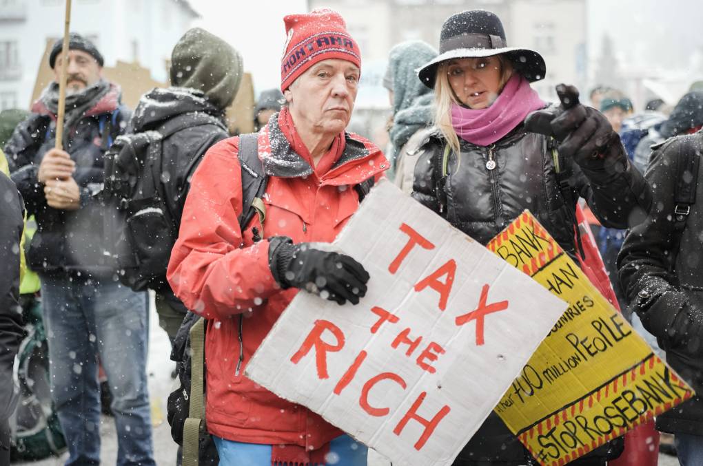 two older people stand in the snow, with signs that say 'tax the rich' in a crowd of protestors