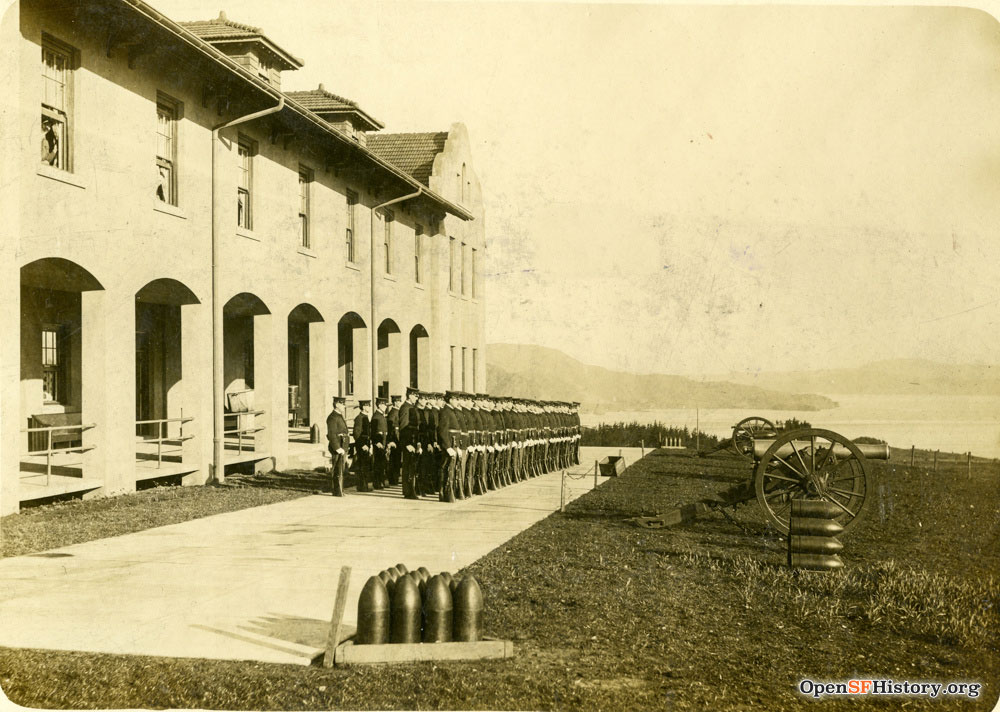 Sepia colored photo shows a large barracks building with three straight rows of soldiers lined up in front. In the distance is water and hills.