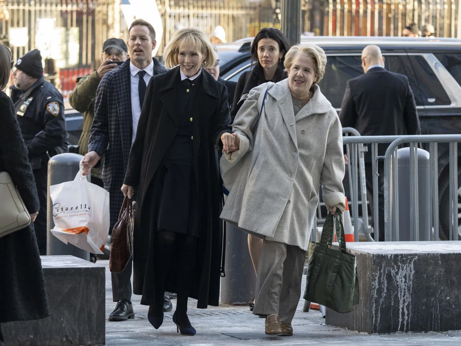 Two white older women in the foreground smile arm in arm as they walk on a sidewalk, with people behind them on a street.