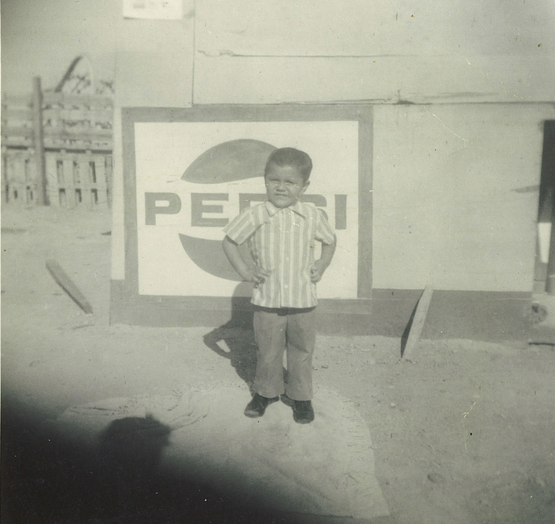 A black and white photo of a 5-year-old boy in front of an old Pepsi logo. The boy stands with a big smile and his hands on his hips.