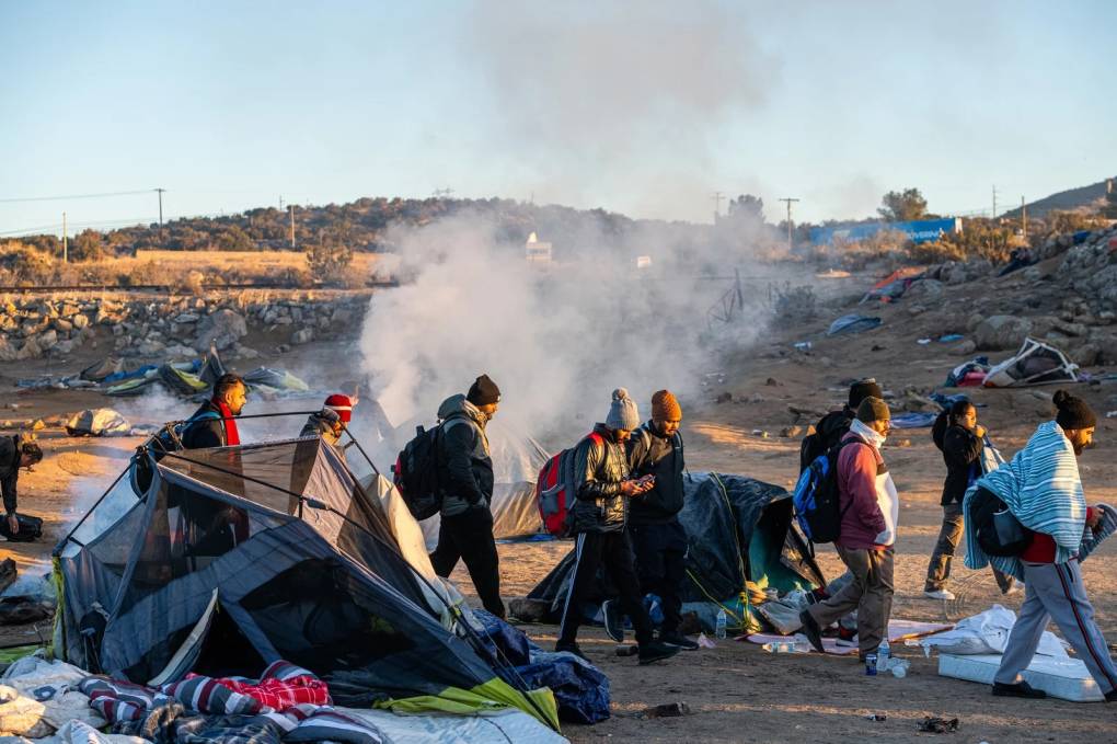 A group of immigrants walking past tents and a smoky fire.