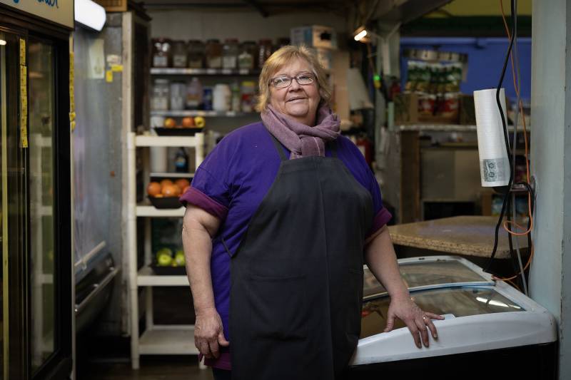 A woman in a purple shirt and a black apron stands in a room with produce in the background