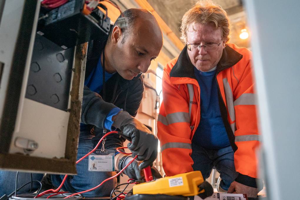 Two men wearing jackets, with the man on the right wearing a neon orange jacket, look down on a yellow device with wiring. The man on the left is putting a wire into the device.