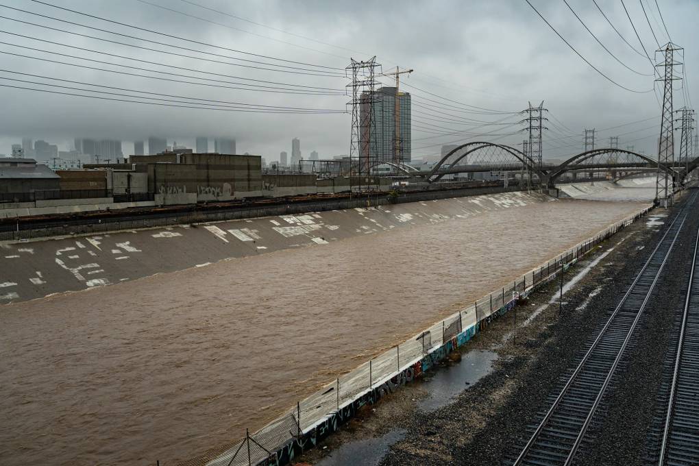 A brown river flows through a city with concrete banks and fencing on either side, buildings and power lines in the backdrop.