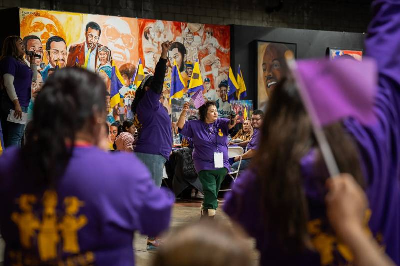 A crowd of people mostly wearing purple shirts and with a mural in the background celebrate and wave flags.