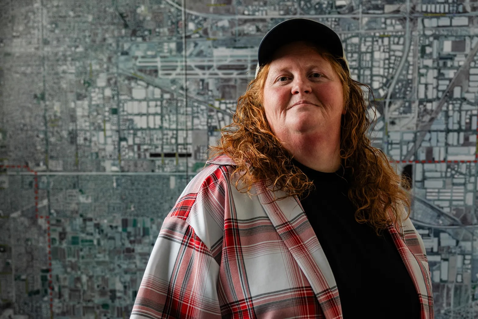 An older white woman with a plaid shirt on a black tshirt and a black truckers hat with a map behind her and looking at the camera with a slight smile. 
