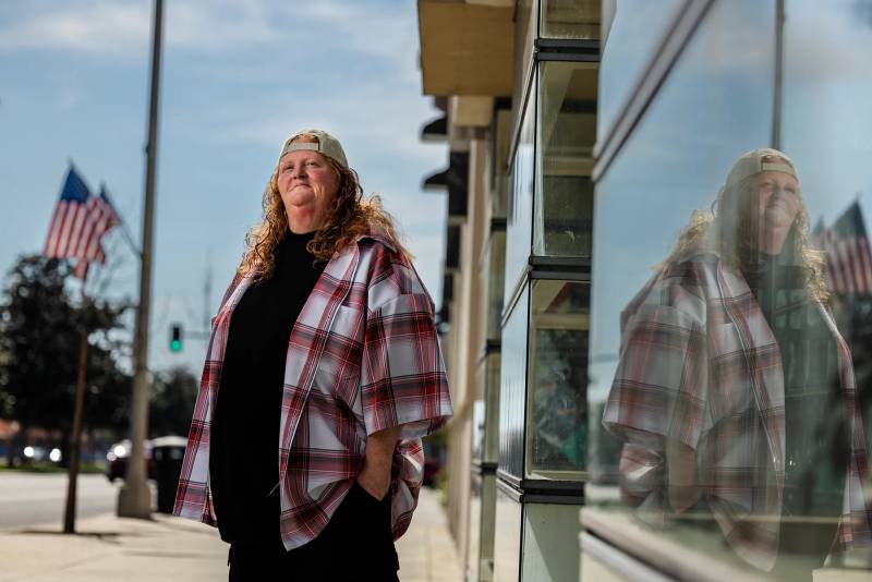An older white woman with a trucker's cap and a plaid shirt stands outside a store on the street, looking away from the camera.