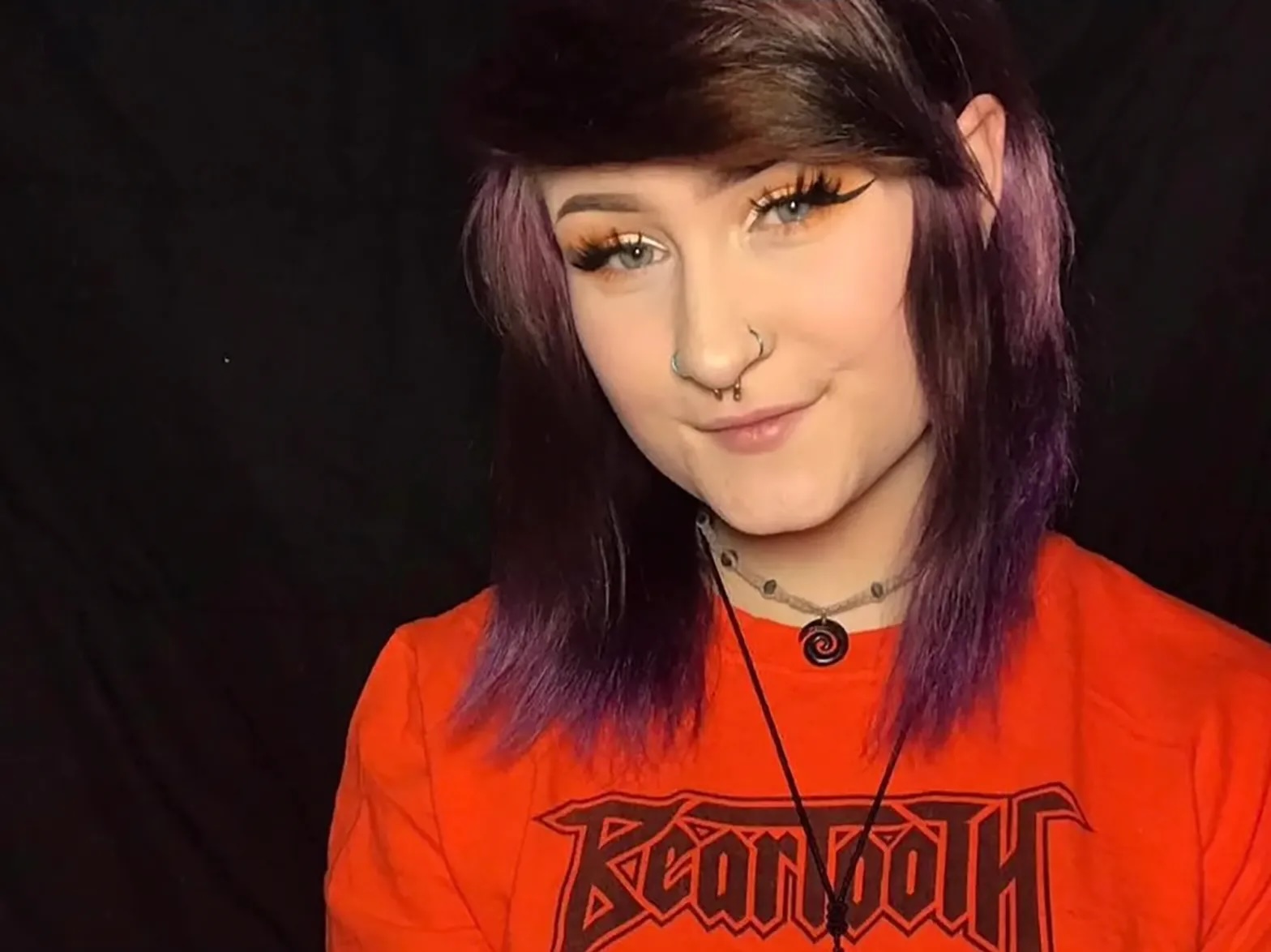A young person with a red shirt eye makeup, shoulder-length hair and necklaces and a nose piercing, smiles at the camera.
