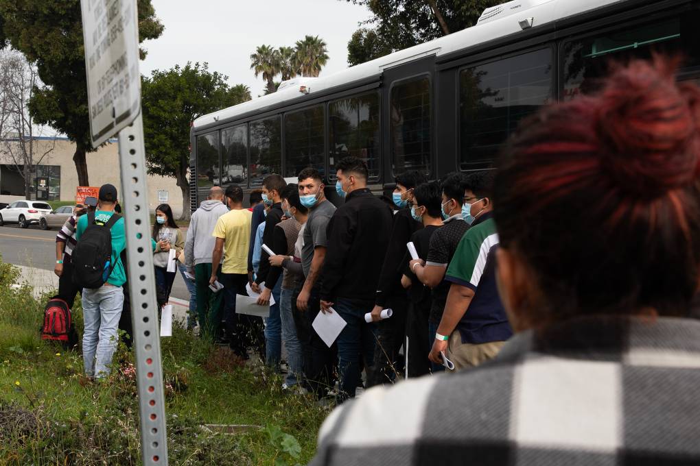 Several people wearing face masks stand in line next to a bus.
