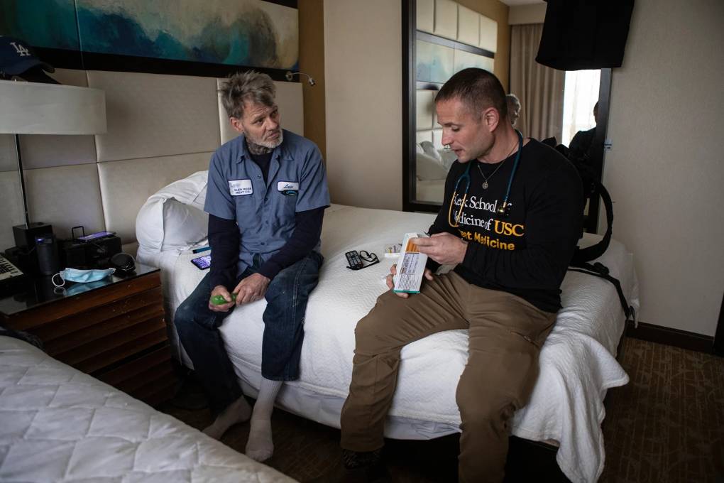 Two men sit on a bed in a hotel room — one with a doctor's stethoscope.