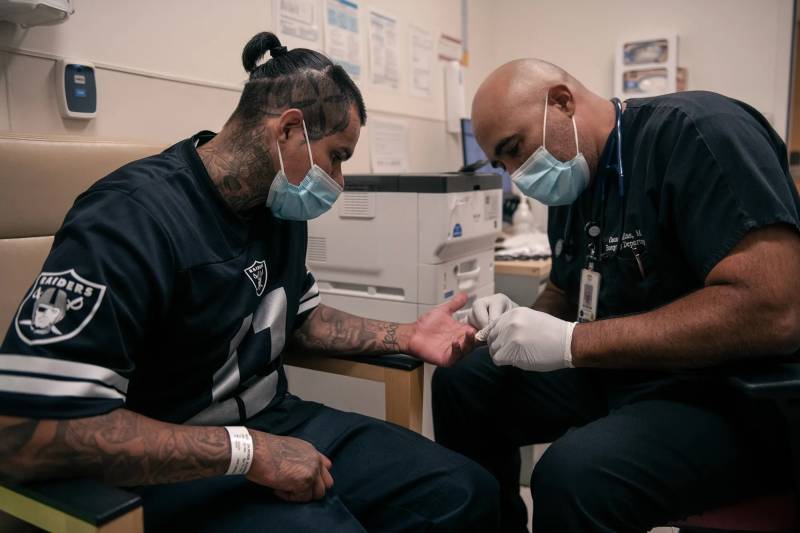 A doctor tends to a patient, both Latino males and wearing masks, in a doctor's office.