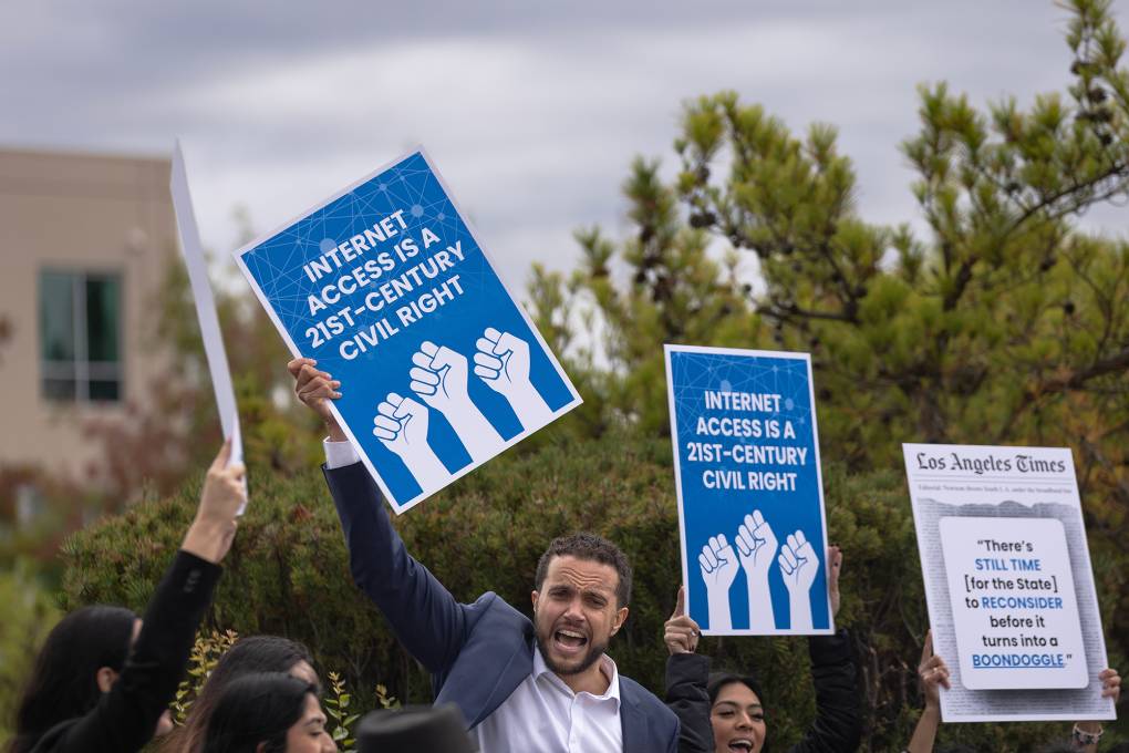 A group of people chant and hold blue signs with fists on them that read "Internet Access is a 21st Century Civil Right."