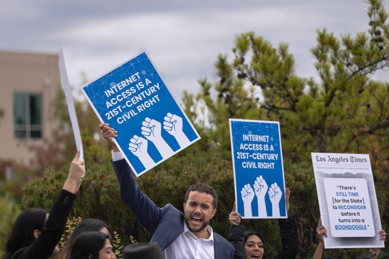 A group of people chant and hold blue signs with fists on them that read "Internet Access is a 21st Century Civil Right."