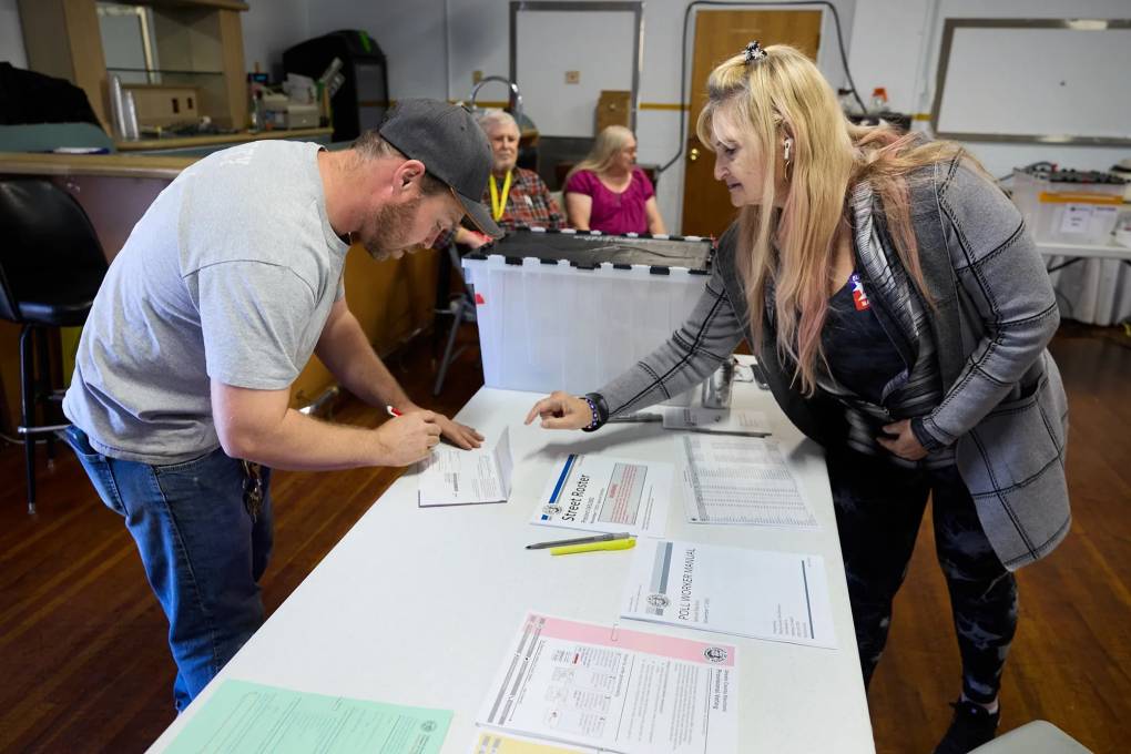 A woman points to a man's ballot as he fills it out. Two other people sit in the background.
