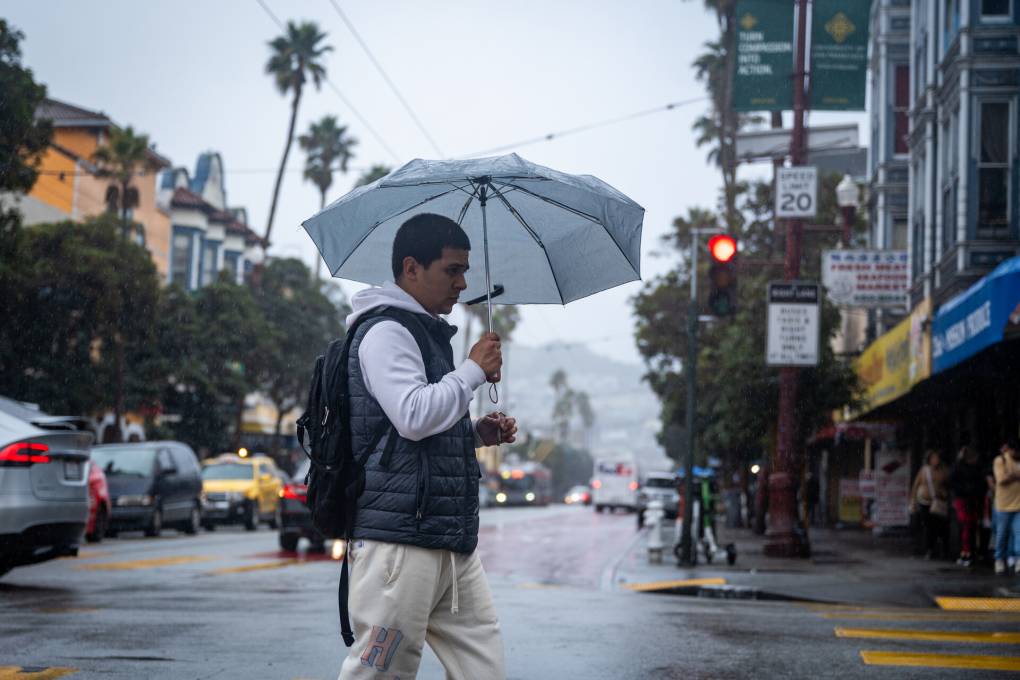 Un hombre lleva puesto varios abrigos y usa un paraguas mientras cruza la calle en una ciudad con muchos carros.