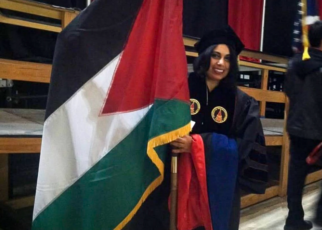 A woman dressed in a graduate gown and standing next to a flag with red, white, black and green.