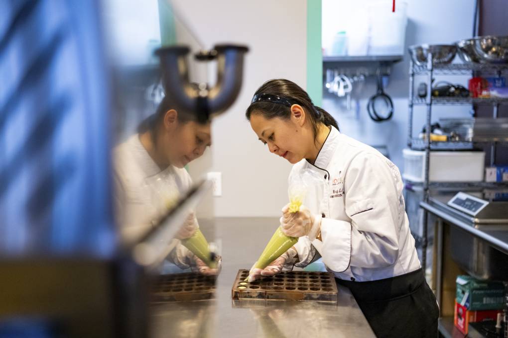 An Asian woman in white shirt leans over a counter working intently on something.