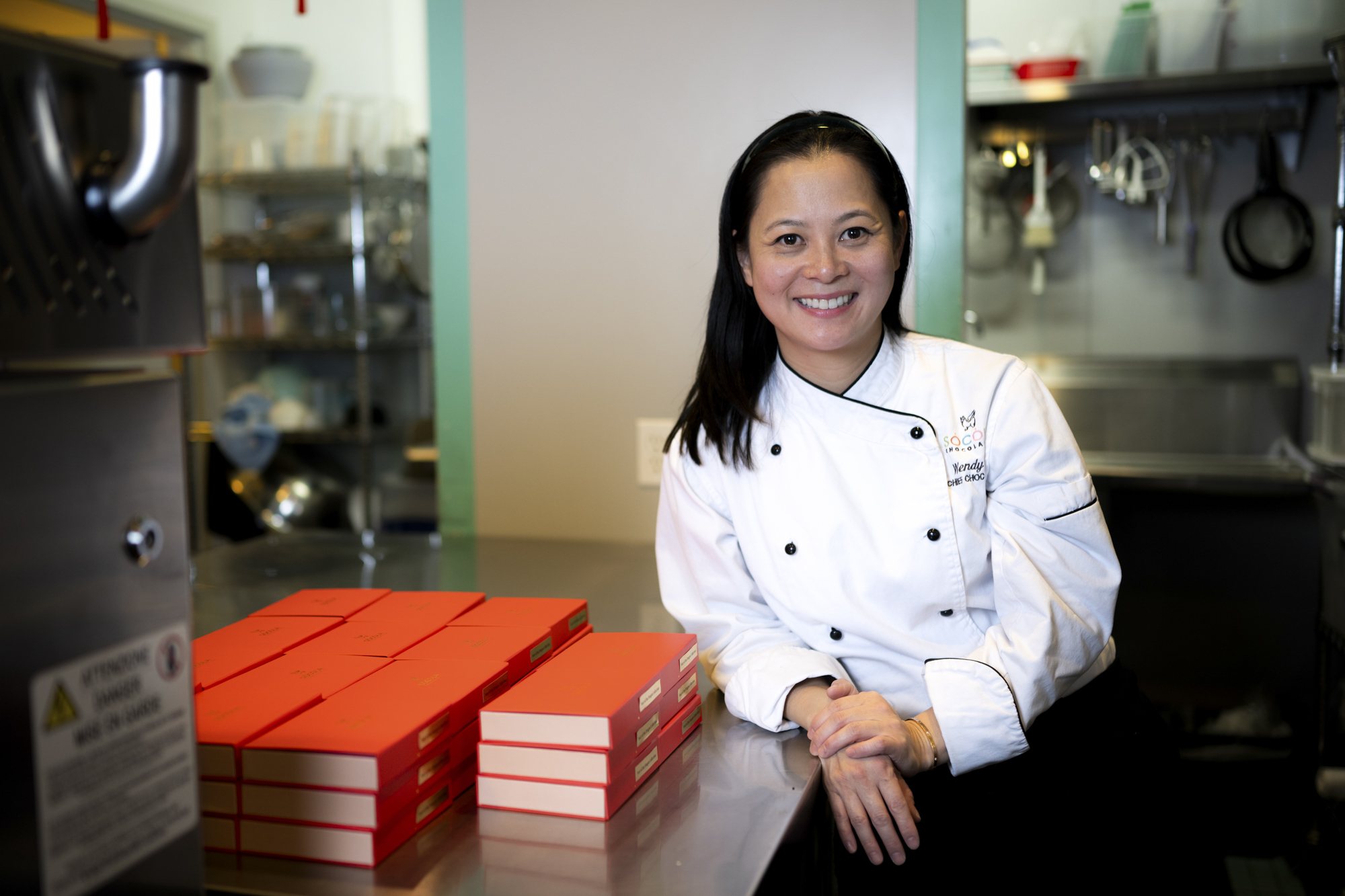 Asian woman in chef's whites leans on a table next to a stack of red boxes.