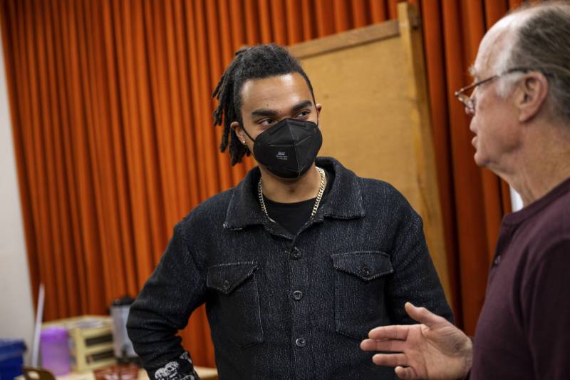 A young Black man with a black face mask speaks to an older white man, with a stage curtain behind them.